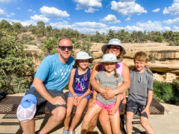 Family of five sitting on a bench overlooking a cliff dwelling at Mesa Verde National Park.